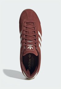 Καστανέρυθρο σουέντ αθλητικό παπούτσι με τρεις λευκές ρίγες και σήμανση εμπορικού σήματος. Στρογγυλερή μύτη, επίπεδος σχεδιασμός με κορδόνια και επένδυση με μαξιλαροειδές εσωτερικό.