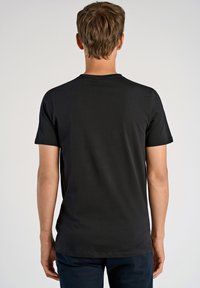 Uomo con capelli castani corti che indossa una t-shirt nera a maniche corte semplice e pantaloni scuri, in piedi di spalle su uno sfondo neutro.