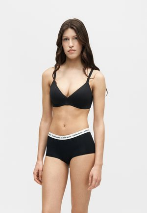 Mladá žena s dlhými hnedými vlasmi, oblečená v čiernej podprsenke a čiernych boyshort nohavičkách s bielym pásikom s logom Björn Borg, stojaca pred jednofarebným pozadím.