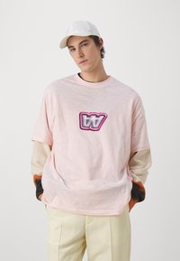 Una camiseta de manga corta de color rosa claro presenta un logo atrevido y multicolor en el pecho. Se lleva con pantalones de color crema y una gorra blanca.