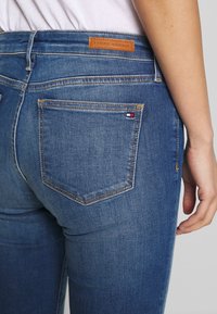 Jeans in denim blu scuro con un'etichetta in pelle marrone "TOMMY HILFIGER", dotati di due tasche posteriori e dettagli di cucitura in vista.