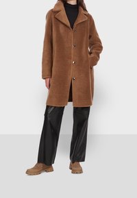 Manteau en polaire marron avec un col cranté, des boutons sur le devant et des poches latérales, associé à un pantalon en cuir synthétique noir et des baskets épaisses couleur beige.