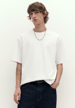 Jonge persoon met halflang haar, gekleed in een eenvoudige witte oversized T-shirt, donkere spijkerbroek, ketting aan de hals en zichtbare tatoeages op de arm.