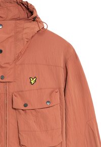 Veste légère orange en tissu texturé, avec capuche réglable et deux poches à rabat à l'avant. Accent de logo jaune sur la poitrine.