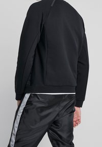 Svart sweatshirt med rund halsringning, stretchtyg och ribbade muddar. Matchas med glänsande svarta byxor med en silverfärgad sidostripa.