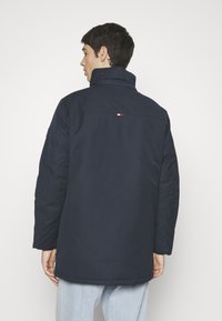 Navy blue insulated parka with a high collar, featuring a small red, white, and blue logo at the back. Smooth, durable fabric.