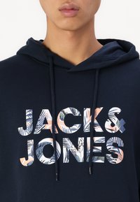 Marineblauer Hoodie mit Kordelzug, der großen "JACK & JONES"-Schriftzug in mehrfarbigen Blumenmustern zeigt. Baumwollstoff mit glatter Textur.