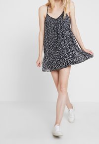 Schwarzes ärmelloses Kleid mit weißem Blumenmuster, fließender Stoff, V-Ausschnitt und gerafftem Vorderteil, kombiniert mit weißen Sneakers.