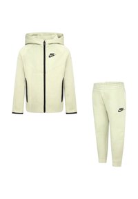 Felpa con cappuccio Nike beige chiaro con zip frontale, cappuccio attaccato e dettagli neri; abbinata a pantaloni coordinati con cinturino elastico in vita e logo.