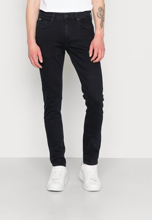 Hombre que lleva vaqueros negros ajustados, zapatillas blancas y una camisa blanca, de pie frente a un fondo gris claro con un tatuaje de serpiente visible en su brazo.