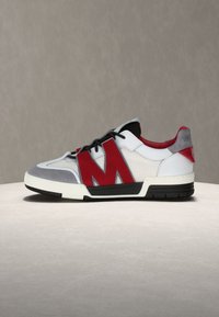 Λευκό και γκρι χαμηλό αθλητικό παπούτσι με μαύρα κορδόνια, κόκκινη λεπτομέρεια "M" στο πλάι, κόκκινη εσωτερική επένδυση και μαύρη και λευκή σόλα σε ουδέτερο φόντο.
