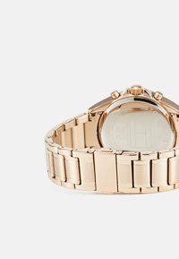 Montre en métal rose gold avec un bracelet à maillons. Présente un boîtier rond, une couronne et un fond en verre transparent montrant le mécanisme interne.