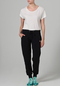 Camiseta blanca de manga corta combinada con pantalones jogger negros con cordón, que presentan un diseño entallado y bolsillos laterales, complementados por zapatillas deportivas turquesas.