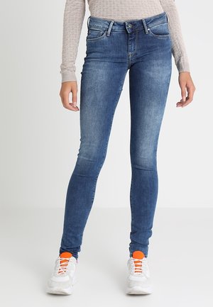 Kvinde iført blå skinny jeans, beige langærmet top og hvide sneakers med klare orange sokker mod en ensfarvet baggrund.