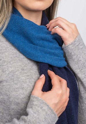 Écharpe en tricot bleu et marine drapée autour du cou. Texture douce avec des teintes alternées, partiellement glissée sous un pull gris. Effet de drapé notable.