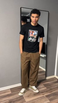 Jauns vīrietis stāv ar rokām kabatās, valkājot melnu grafisko t-kreklu, brūnas bikses un zaļus sporta apavus pie pilna garuma spoguļa.