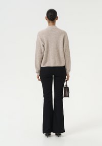 Beige gebreide trui met een relaxte pasvorm, gecombineerd met zwarte flared jeans. Model houdt een kleine donkerhandtas vast, gezien vanaf de achterkant.