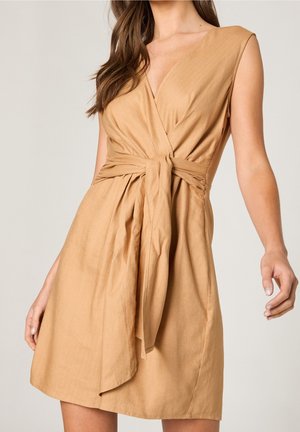 Beżowa, bezrękawnikowa sukienka kopertowa z głębokim dekoltem w serek i wiązanym paskiem w talii, noszona przez kobietę o długich brązowych włosach na jednolitym tle.