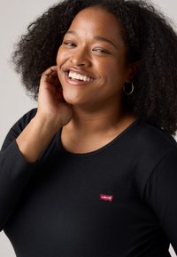 Top negro de manga larga con canales, escote redondo y un pequeño parche de logotipo rojo de Levi's en el pecho. La modelo tiene el cabello rizado y lleva un pendiente de aro dorado.