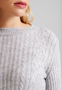 Ljusgrå kabelstickad tröja av mjukt material, med ribbad halsringning och texturerade mönster över tyget.