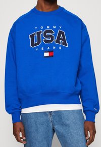 Felpa in cotone blu con la scritta "TOMMY USA JEANS", dotata di un piccolo logo applicato. Maniche e orlo a coste, indossata sopra una camicia bianca.