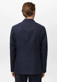 Granatowy blezer wykonany z gładkiego materiału; ma wyprofilowane ramiona, klapy w kształcie notcha oraz jedno rozcięcie z tyłu. Minimalistyczny design.