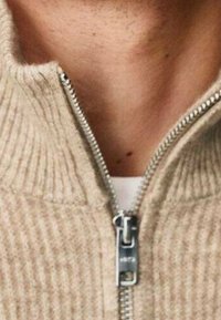 Maglione beige a coste parzialmente zipato, che mostra la pelle e una maglietta bianca sotto il colletto.