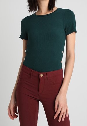 Haut court à rayures vert foncé avec des accents de boutons dorés sur le côté, associé à un jean bourgogne ajusté. Texture lisse et couleurs unies.