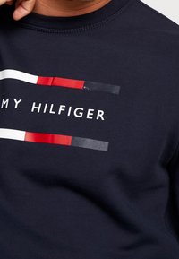 T-shirt in cotone blu navy con un logo bianco e strisce orizzontali in rosso, bianco e grigio sul davanti. Texture liscia con colletto a girocollo.