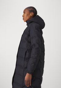 Lungo cappotto trapuntato nero con cappuccio, caratterizzato da un design a quadrati e una superficie liscia. Include chiusure a bottone e tasche laterali.