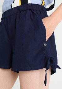 Marinblå shorts med resårmidja, sidofickor och dekorativa snören vid fållen, tillverkade av ett lättviktigt tyg.