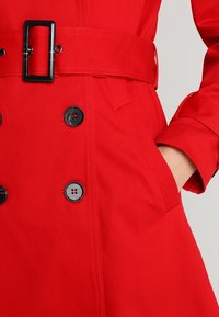Červený trenčkot s páskem, černými knoflíky, hladkým texture a bočními kapsami. Design obsahuje dvouřadé zapínání a široké manžety.