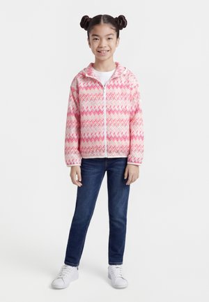 Νεαρή κοπέλα όρθια, φοράει ροζ και λευκό φούτερ με φερμουάρ και σχέδιο ζιγκ ζαγκ, σκούρο μπλε τζιν και λευκά αθλητικά παπούτσια, χαμογελώντας στην κάμερα.