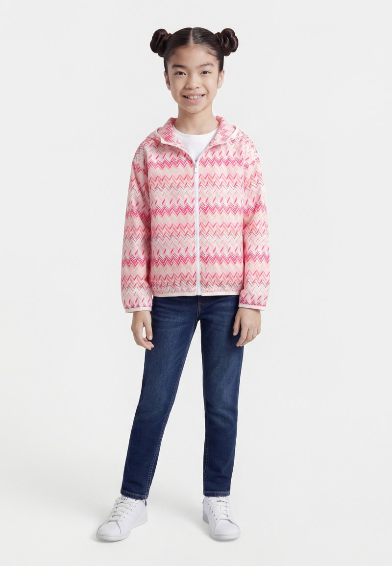 Jeune fille debout, portant une veste zippée à motifs zigzag roses et blancs, un jean bleu foncé et des baskets blanches, souriant à la caméra.