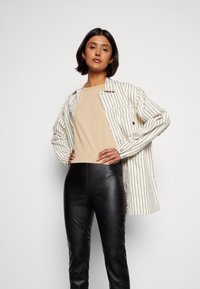 Casaco creme riscado sobre uma blusa bege e calças de pele pretas. O casaco apresenta um bolso no peito e um corte descontraído; texturas suaves em toda a peça.