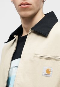 Artimos plano asmuo dėvi smėlio spalvos Carhartt striukę su juodu apykakle ir po ja juodą marškinėlį su grafiniu raštu.