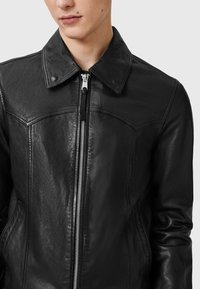 Zwarte leren jas met een puntkraag, ritssluiting met metalen tanden en een getailleerde pasvorm. Gladde textuur met minimale details.