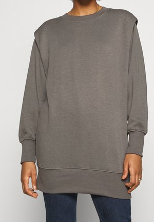 Person iført en løs, langærmet, grå sweatshirt med ribbede manchetter og kant, kombineret med mørkeblå jeans, stående foran en ensfarvet baggrund.