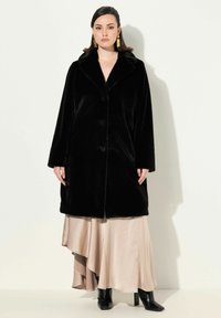 Abrigo de piel sintética negro con cuello de solapa, que presenta grandes botones y mangas largas. Llevado sobre un vestido beige fluido y botas negras hasta el tobillo.