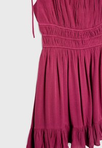 Robe rose sans manches avec taille élastique froncée, corsage plissé et ourlet à volants, suspendue sur un fond uni.