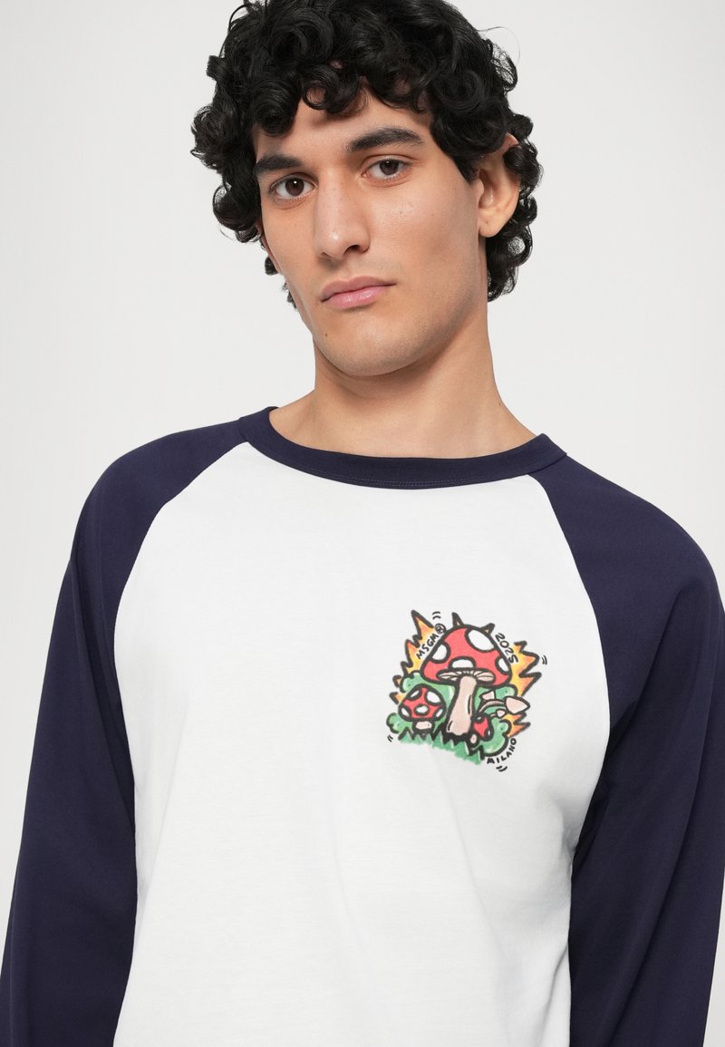 Chemise à manches longues blanche avec des manches raglan bleu marine, décorée d'un graphique coloré de champignon sur la poitrine. Tissu lisse avec une coupe décontractée.