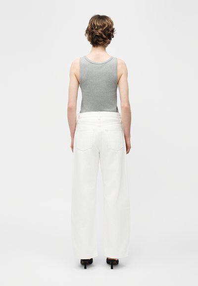 Άτομο με κοντά σγουρά μαλλιά φοράει γκρι αμάνικη μπλούζα, λευκό παντελόνι με φαρδιά πόδια και μαύρα παπούτσια με τακούνι, γυρισμένο προς τα πίσω σε λευκό φόντο.