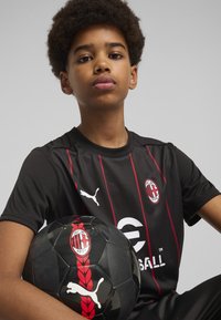 Zwart voetbalshirt met rode verticale strepen, met een rond embleem. De jongen houdt een zwarte voetbal vast met rode details en logo.
