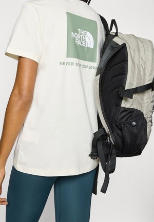 Osoba nosząca białą koszulkę The North Face i turkusowe legginsy, niosąca szaro-czarny plecak na jednym ramieniu.