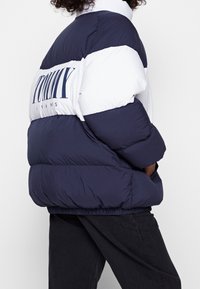 Person som bär en marinblå och vit puffjacka med stor "TOMMY JEANS" text på ryggen, i kombination med svarta byxor.