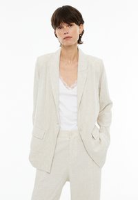 Light beige linen blazer with a notched lapel, two front pockets, and two buttons, paired with a white tank top featuring lace trim.