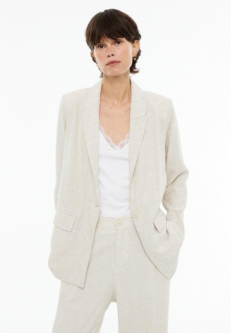 Light beige linen blazer with a notched lapel, two front pockets, and two buttons, paired with a white tank top featuring lace trim.