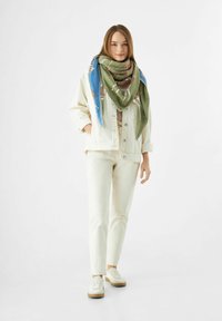 Grande écharpe verte et bleue à motif portée sur une veste crème et un pantalon, dotée d'une coupe ample et de baskets décontractées avec une semelle légère.