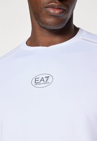 Valge võrkmaterjalist sportpoliis, millel on ümar kaelus ja rinnal must "EA7 Emporio Armani" logo. Silkus tekstuur, lühikesed varrukad.