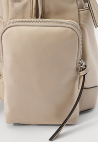 Zaino in nylon beige con una tasca frontale con zip, tessuto texturizzato e dettagli delle zip in contrasto. Include una piccola maniglia per un accesso facile.